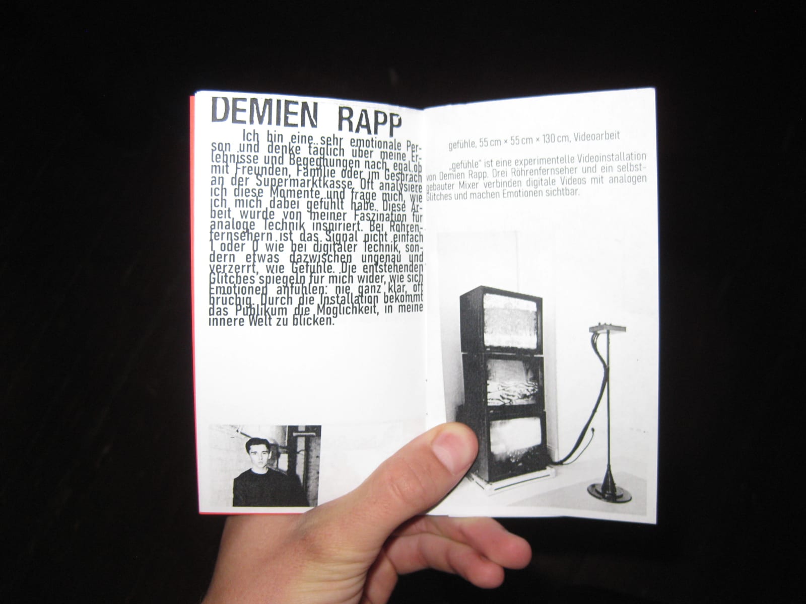 A hand holds the exhibition zine open at the page about Demien Rapp, with a photo of the stacked televisions.