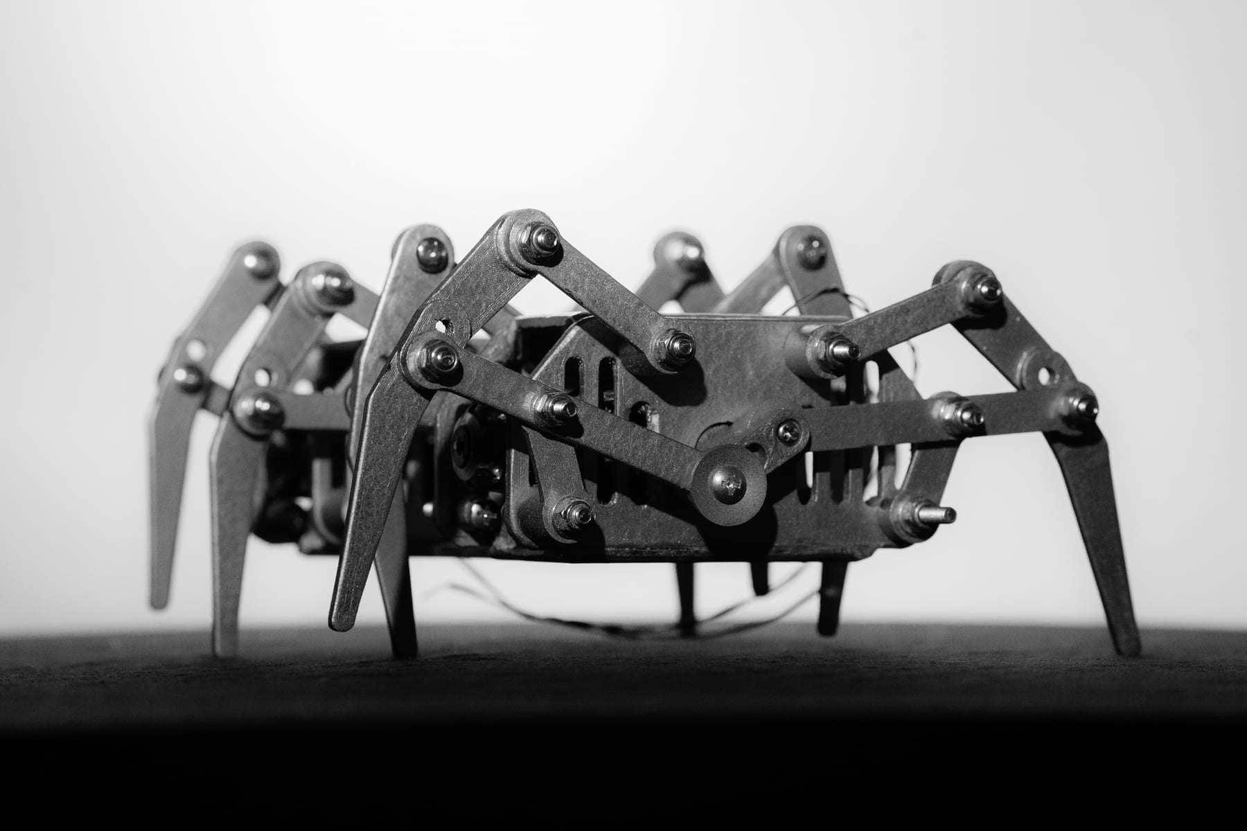 mine: ein schwarz-weißes Foto eines niedrigen, spinnenartigen Laufroboters aus 3D-gedruckten Gestängen auf einem dunklen Tisch, von Hand metallisch lackiert und mit einem dünnen Kabel gefesselt.