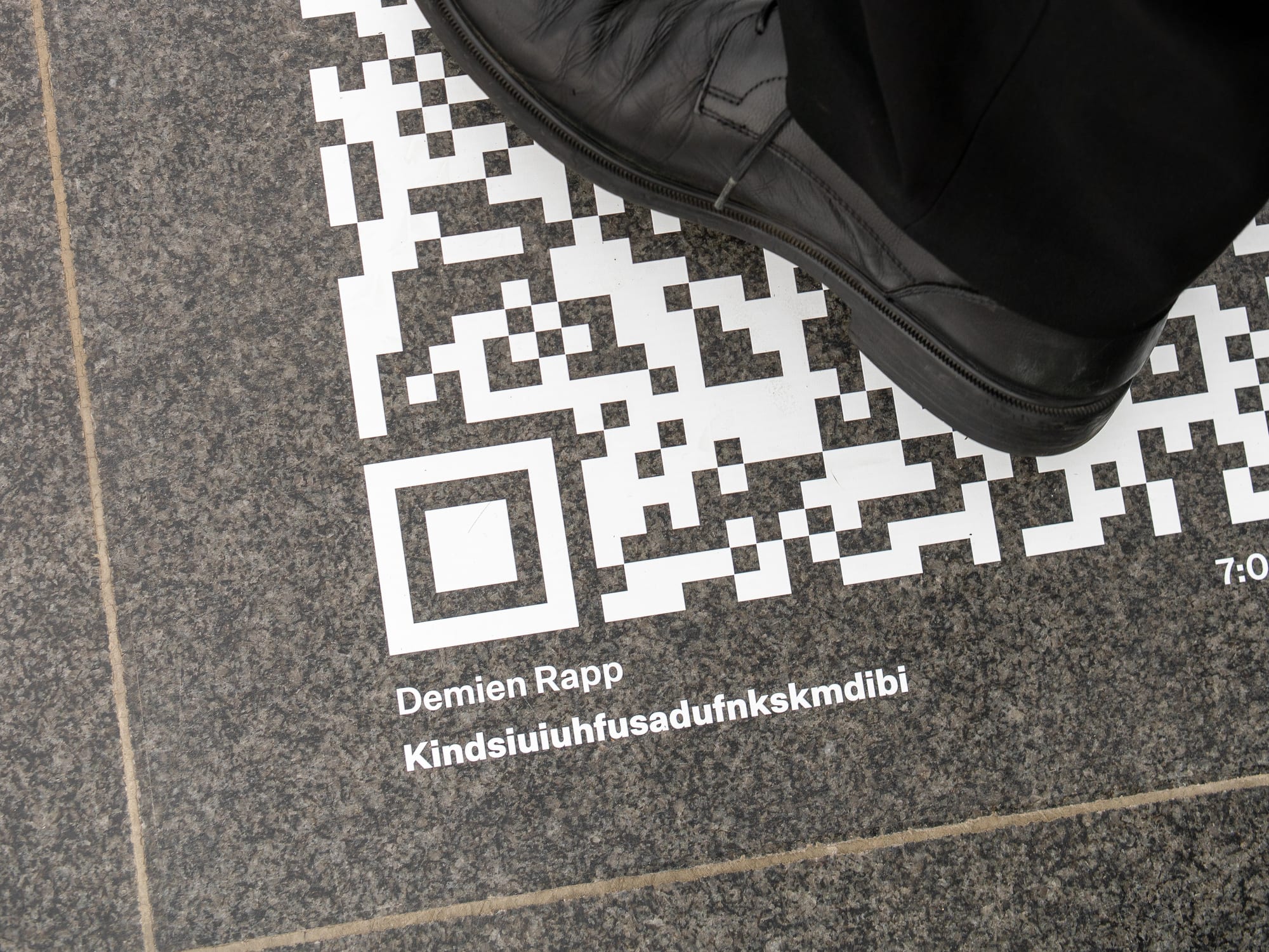 A QR code printed on the stone floor at K21, labelled Demien Rapp, Kindsiuiuhfusadufnkskmdibi, 7:00, a visitor’s shoe at its edge.