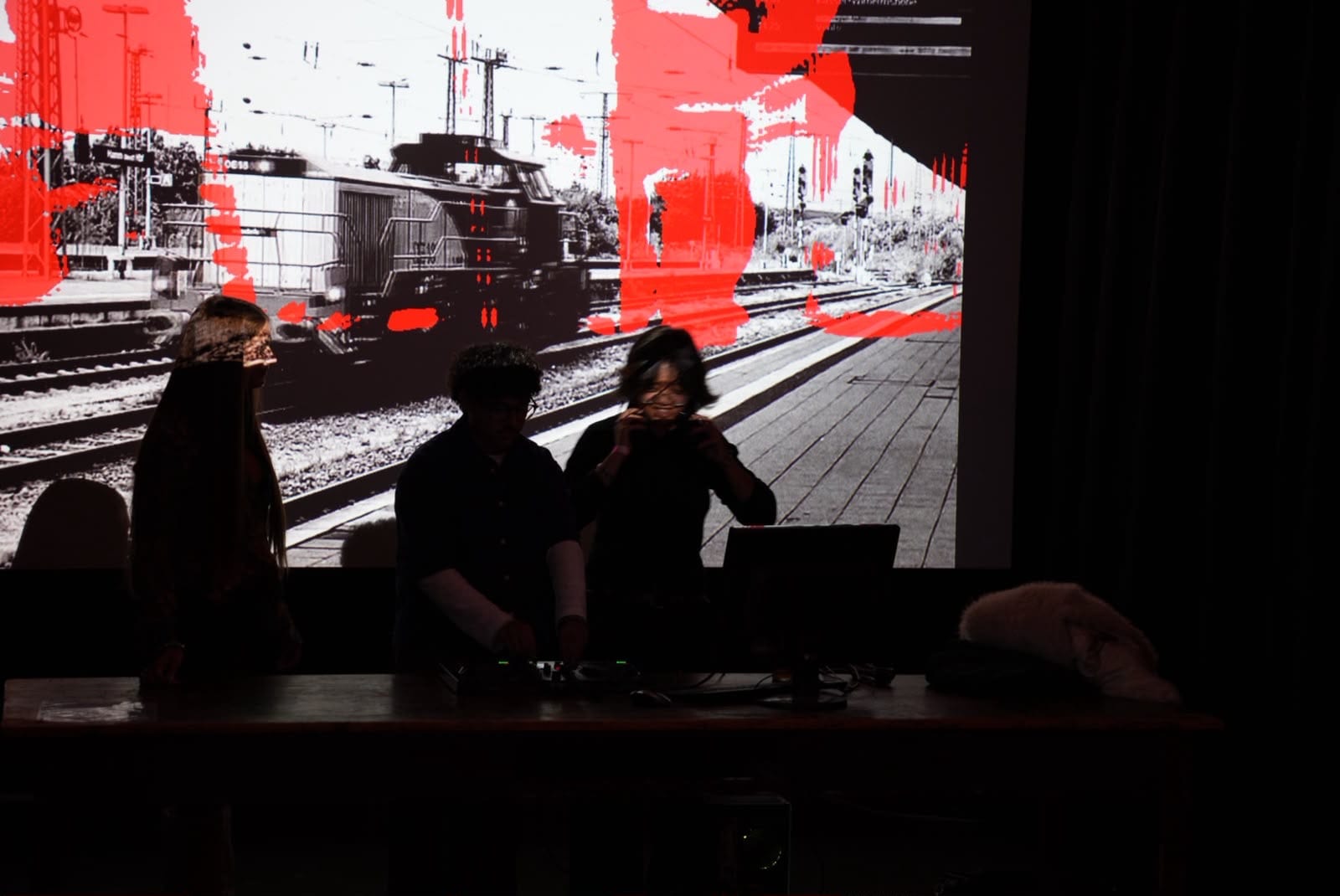The video installation: a black-and-white station scene projected with red artefacts where people moved, three people at the desk in front of it.